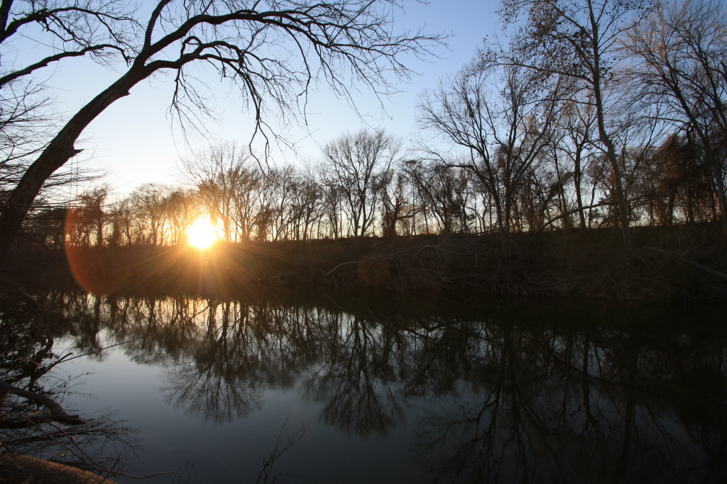 Sun setting through silhouettes of bare winter trees reflected in a calm river.