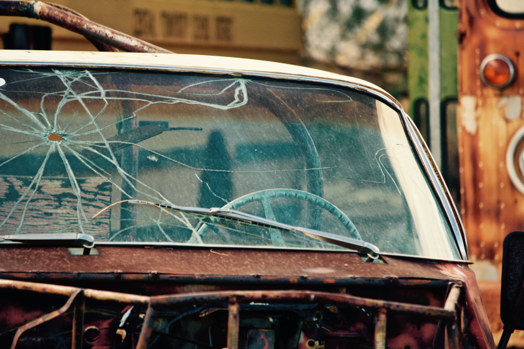 A hole in a rusted out car's windshield has created a spider crack through the glass as it sits deserted in a junkyard